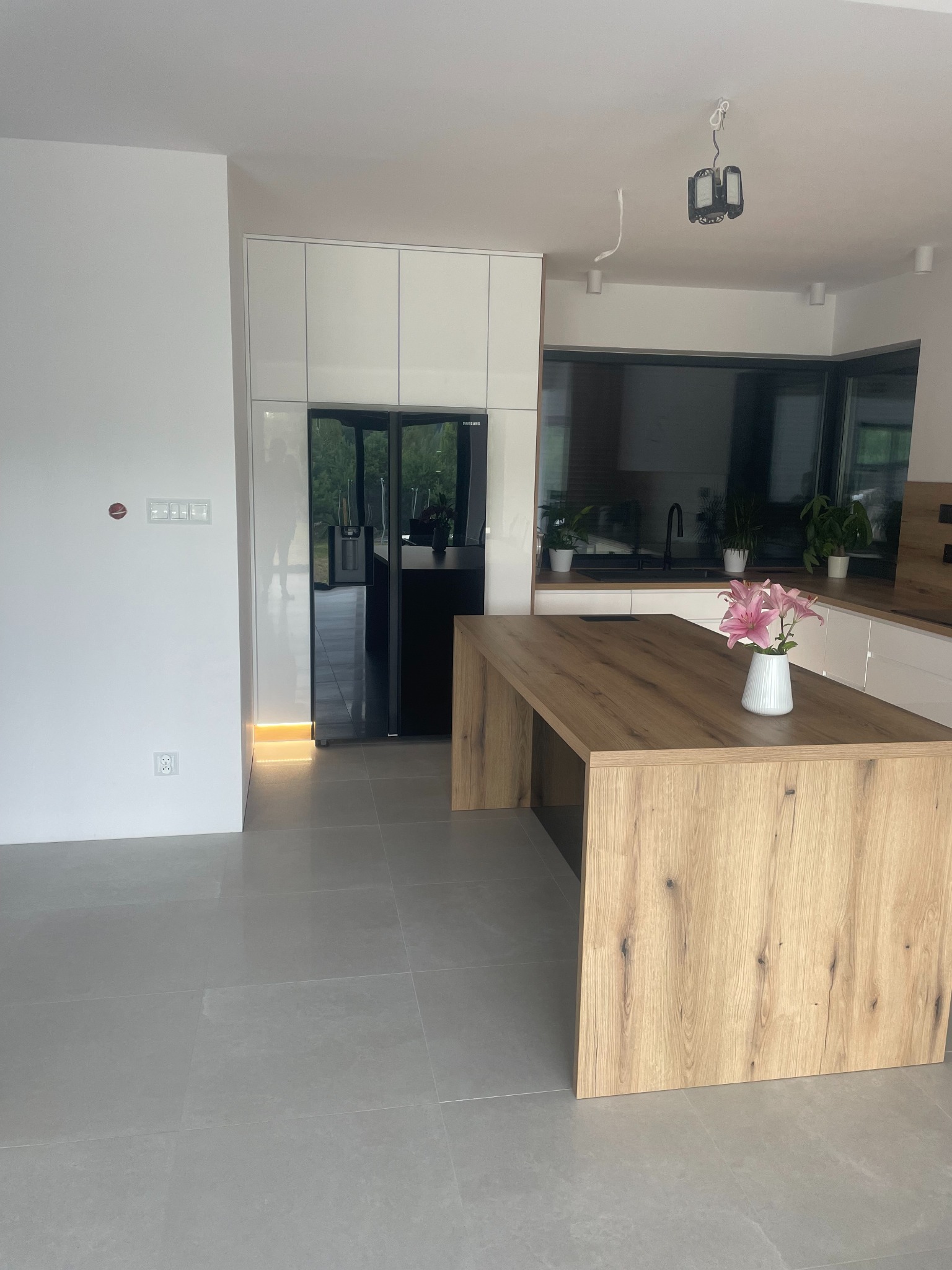 Nowoczesna kuchnia z wyspą z blatem imitującym drewno, białymi szafkami i czarną lodówką. Na wyspie stoi biały wazon z różowymi liliami.