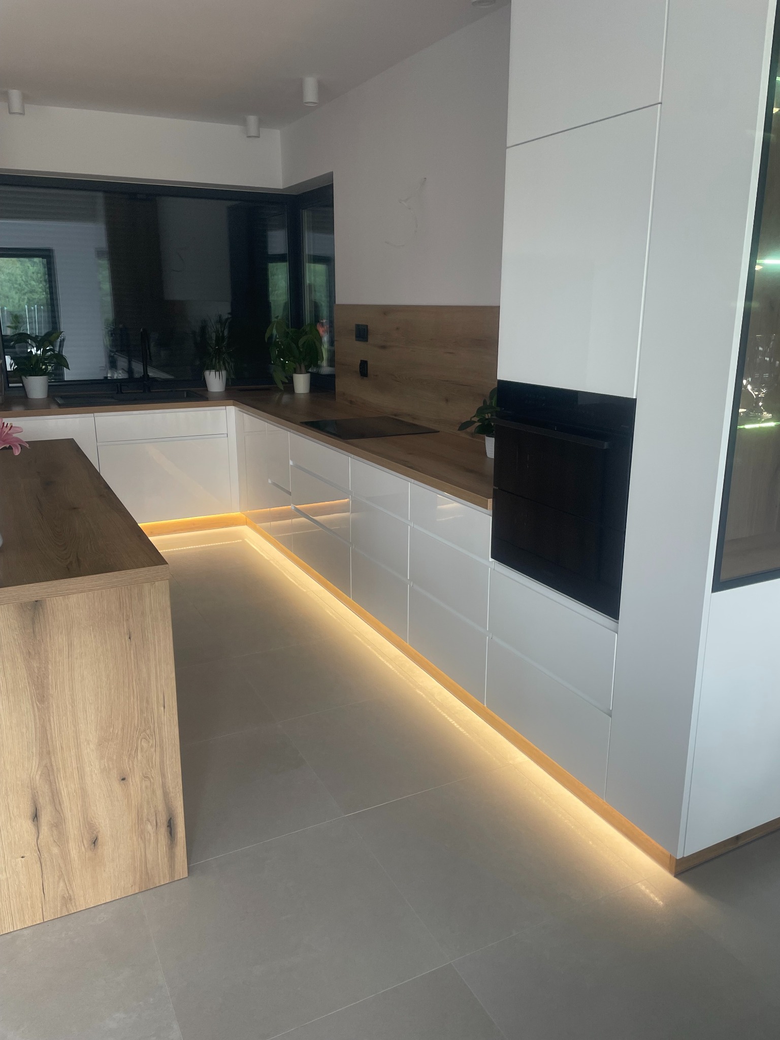 Nowoczesna kuchnia z białymi frontami, drewnianym blatem i podświetleniem LED wzdłuż dolnych szafek, widok na wyspę kuchenną i duże okno.