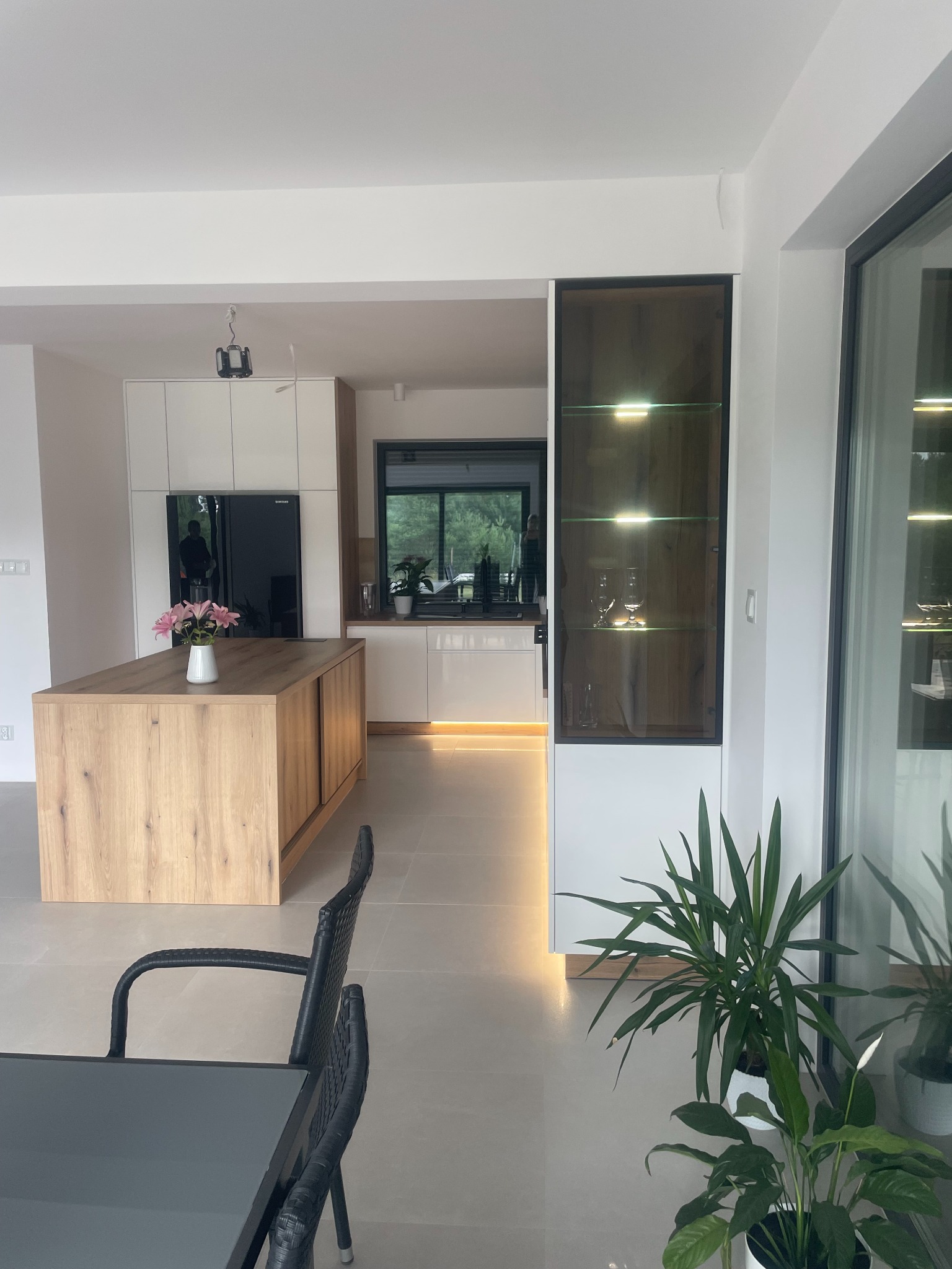 Nowoczesna kuchnia z wyspą o drewnianym wykończeniu, białymi szafkami i oświetleniem LED pod szafkami, widok z jadalni z czarnymi krzesłami.