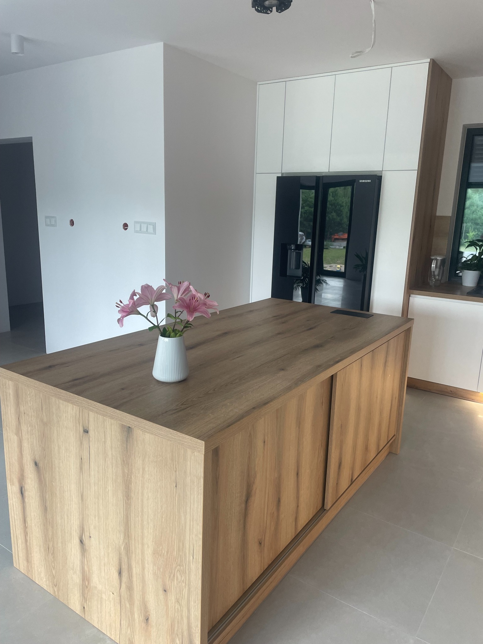 Wyspa kuchenna z blatem o strukturze drewna, z przesuwnymi drzwiami, na której stoi biały wazon z różowymi liliami, w tle zabudowa kuchenna z lodówką.