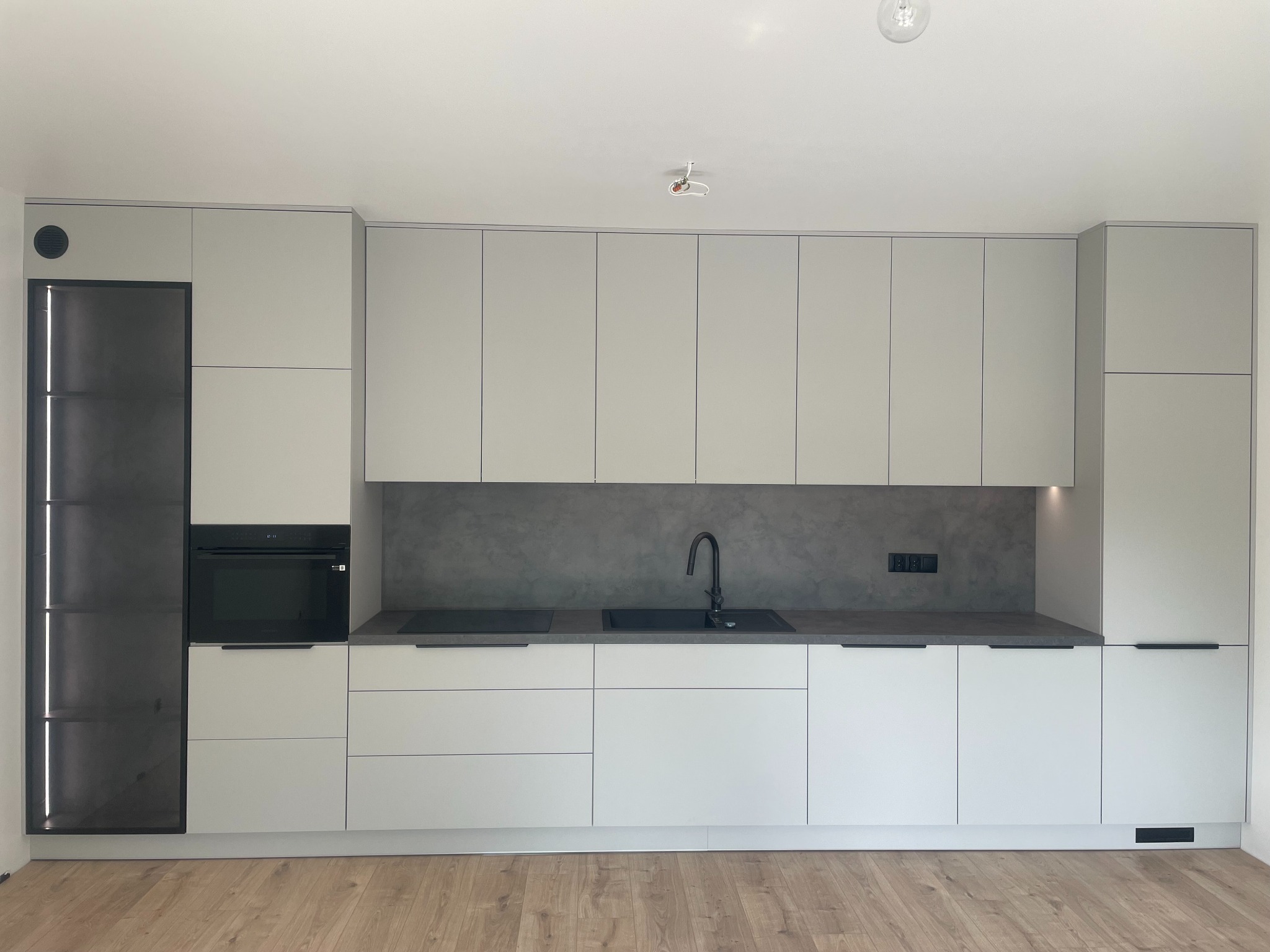 Nowoczesna kuchnia z szarymi frontami, czarną armaturą i szklaną witryną z oświetleniem LED, widok na drewnianą podłogę.