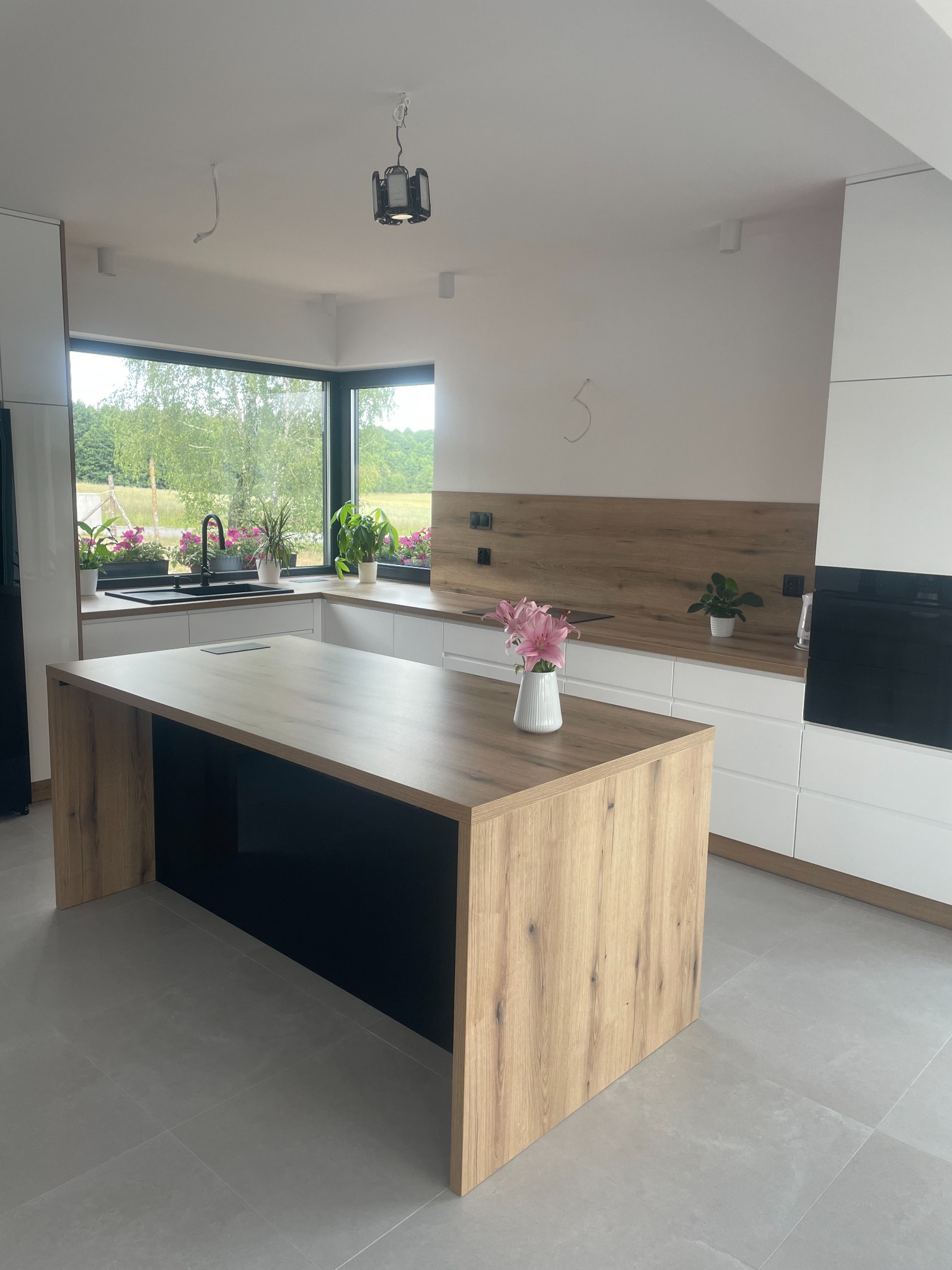 Nowoczesna kuchnia z wyspą o drewnianym blacie i czarnych wnękach, białe szafki, czarny zlew pod oknem z widokiem na zieleń, kwiaty w wazonie na wyspie.