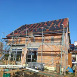 Dom w trakcie budowy z czerwonej cegły, częściowo pokryty dachem z czarnych dachówek, rusztowania wokół budynku, błękitne niebo w tle.