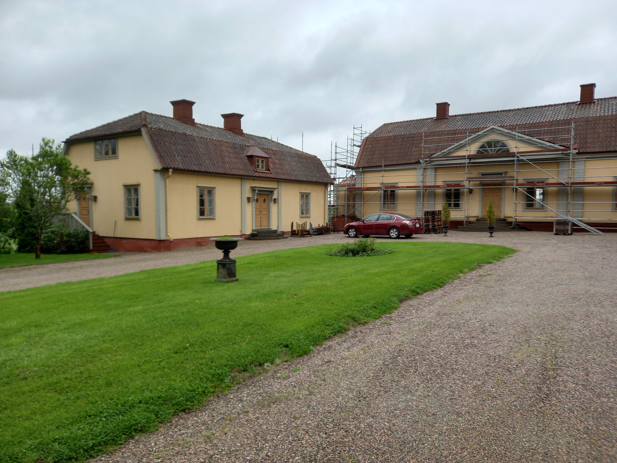 Dwa żółte budynki z czerwoną dachówką, połączone trawnikiem i żwirową drogą, jeden budynek w trakcie prac remontowych z rusztowaniem, czerwony samochód zaparkowany obok.