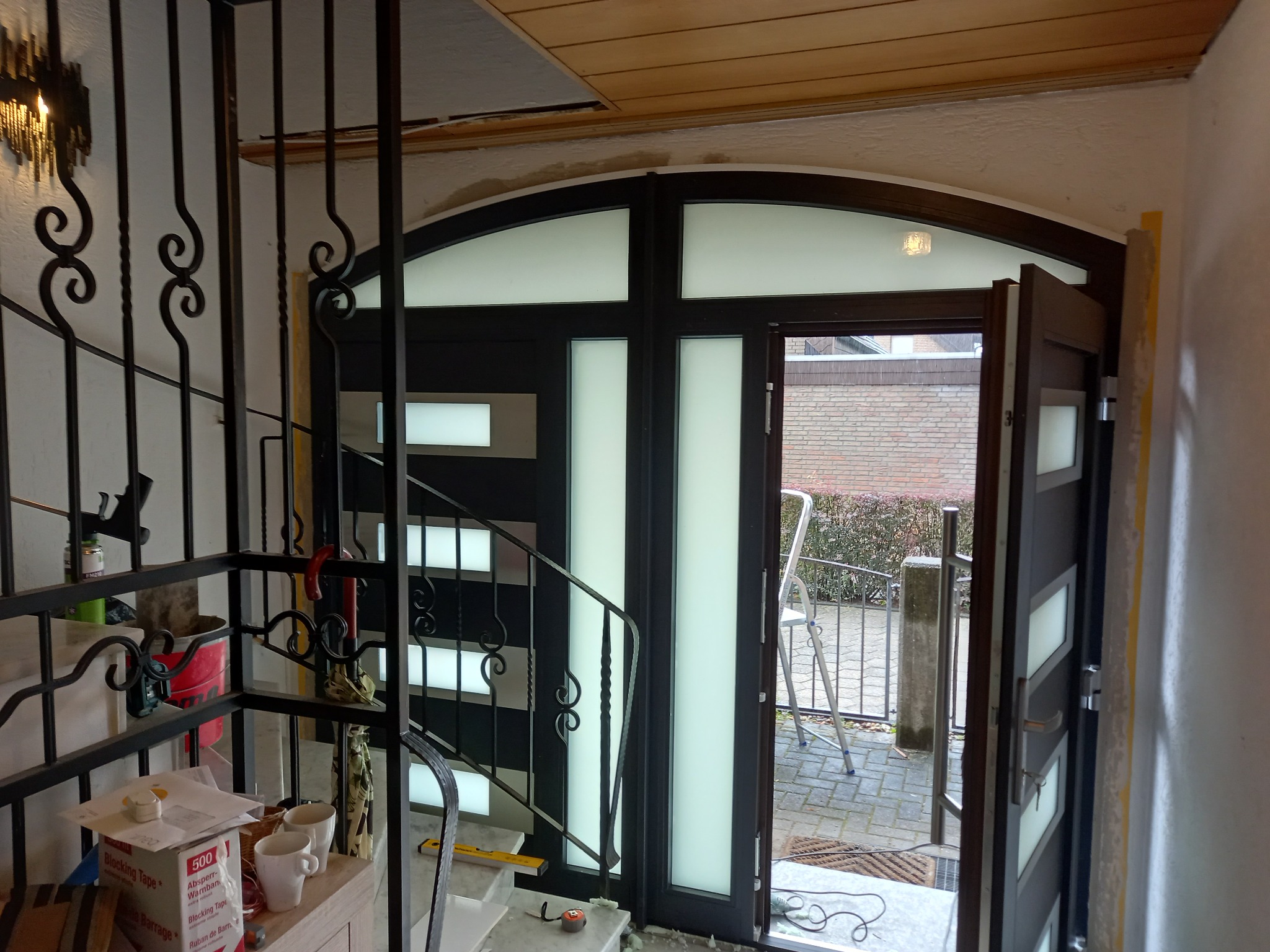 Montaż nowoczesnych, dwuskrzydłowych drzwi wejściowych z przeszkleniami, widoczne schody z ozdobną, kutą balustradą i fragment elewacji zewnętrznej z drabiną.