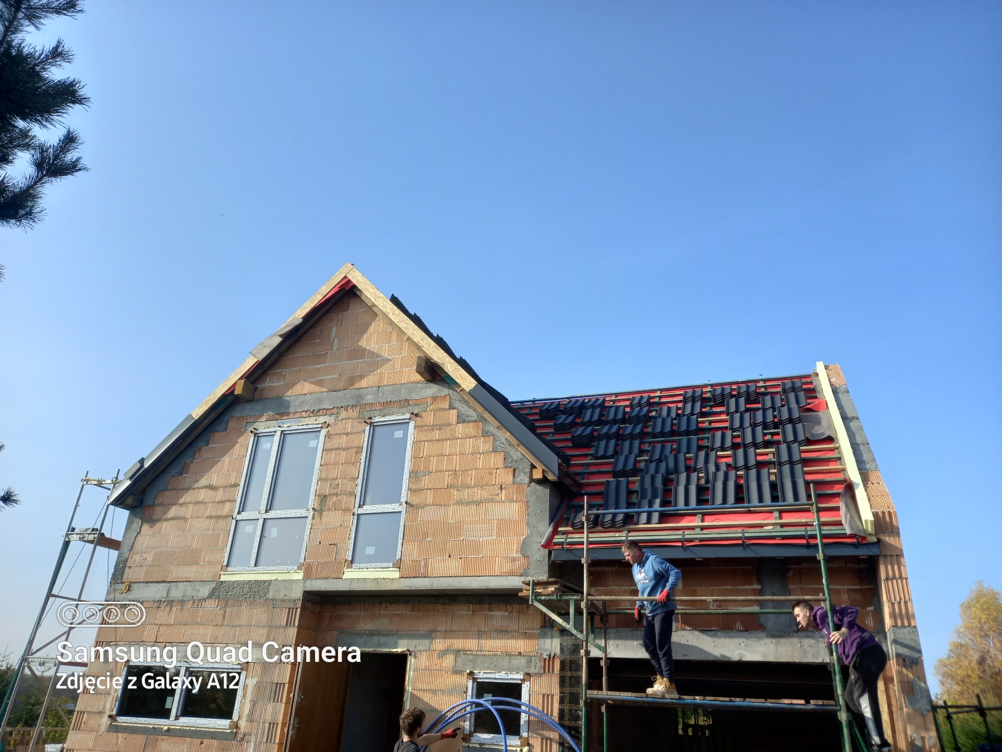 Budowa domu z cegły: widok na dwukondygnacyjny budynek w trakcie wznoszenia, z częściowo ułożonym dachem z czarnej dachówki, widoczne rusztowania z pracownikami na wysokości.