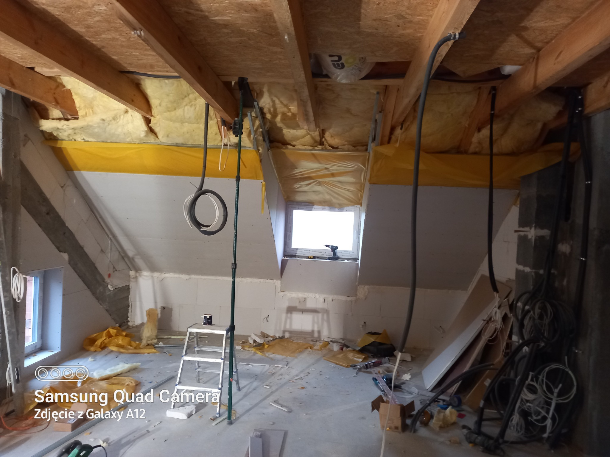 Poddasze w trakcie adaptacji: izolacja wełną mineralną, folia paroizolacyjna, stelaż pod płyty gipsowe, widoczne kable elektryczne, drabina i narzędzia.