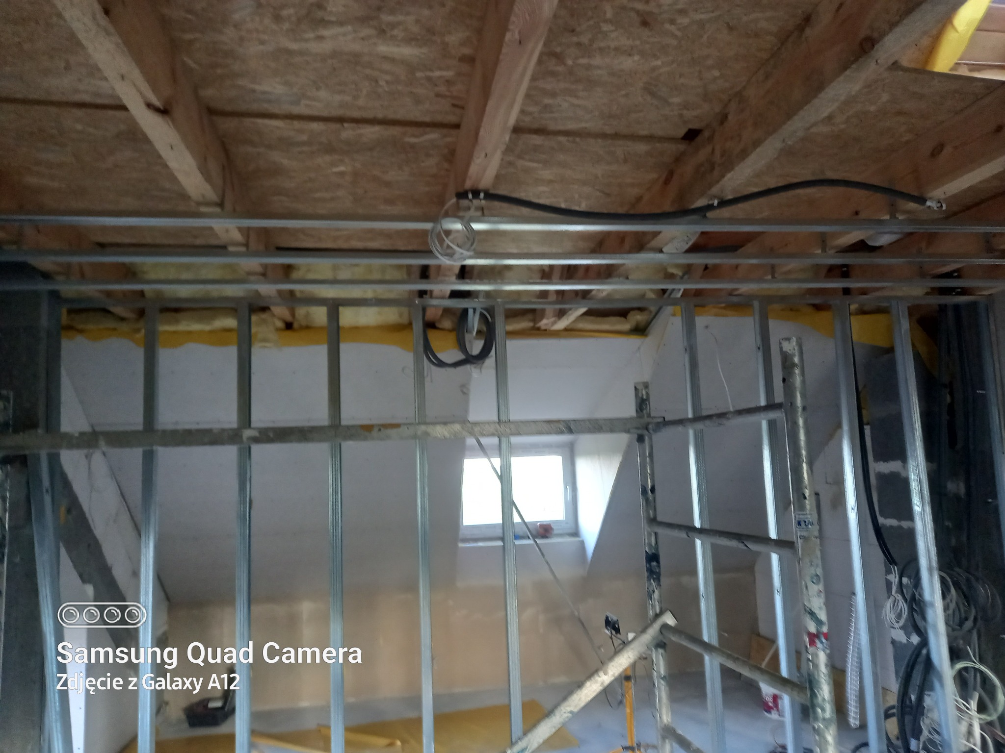 Wnętrze poddasza w trakcie remontu: widoczne metalowe profile ścian działowych, częściowa izolacja z żółtej wełny mineralnej, drewniana konstrukcja dachu i okno na poddaszu.