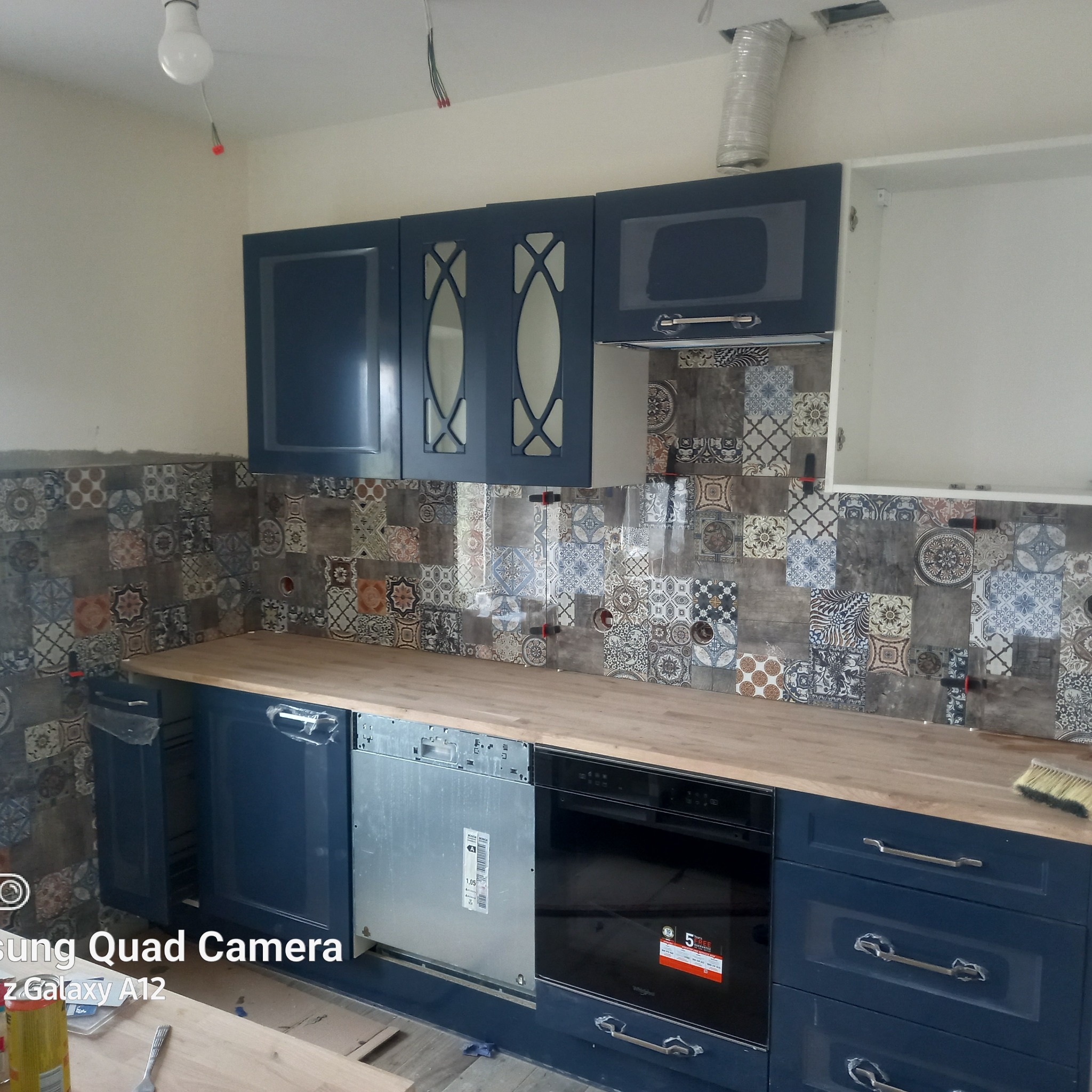 Montaż niebieskiej zabudowy kuchennej z drewnianym blatem i wzorzystymi płytkami backsplash, widoczne niedokończone prace i narzędzia.