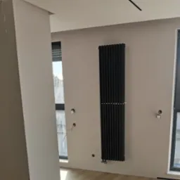 Wnętrze w trakcie prac wykończeniowych: zamontowany czarny, pionowy grzejnik żeberkowy na beżowej ścianie, widoczne otwory na gniazdka elektryczne i przewody, okna z roletami wewnętrznymi, fragment...