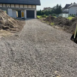 Droga dojazdowa do nowego domu w trakcie budowy, wysypana drobnym, szarym żwirem, obok pryzmy ziemi.