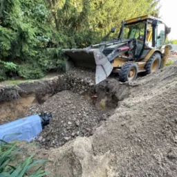 Żółta koparko-ładowarka wysypuje żwir do wykopu z widocznym elementem drenażu w ziemi, na tle zielonych drzew.