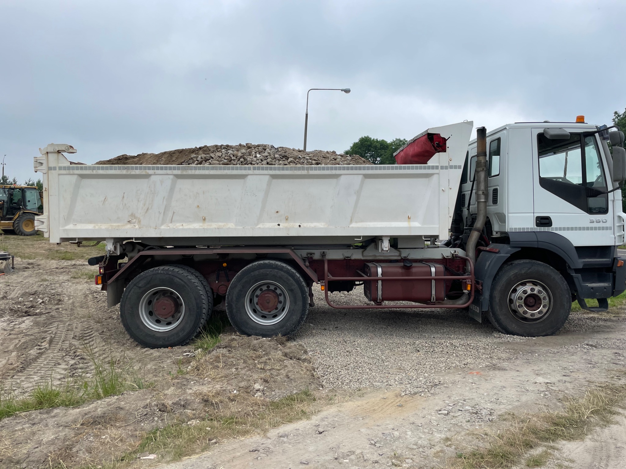 Ciężarówka z wywrotką wypełniona ziemią i gruzem na placu budowy, widok z boku, na pierwszym planie teren z widocznymi śladami opon, w tle słup oświetleniowy.
