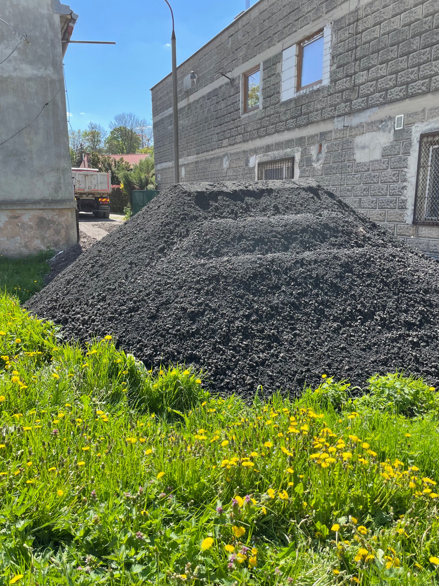 Góra żwiru w Radomiu, składowana na trawie z kwitnącymi mniszkami, w tle budynek mieszkalny z szarej cegły i częściowo widoczna ciężarówka.