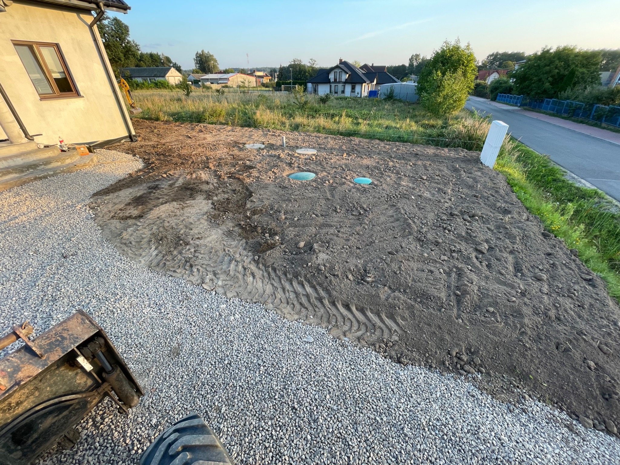 Przygotowany teren pod budowę z widocznymi studzienkami kanalizacyjnymi i częściowo wysypanym żwirem, w tle domy i zieleń, widoczny fragment maszyny budowlanej.