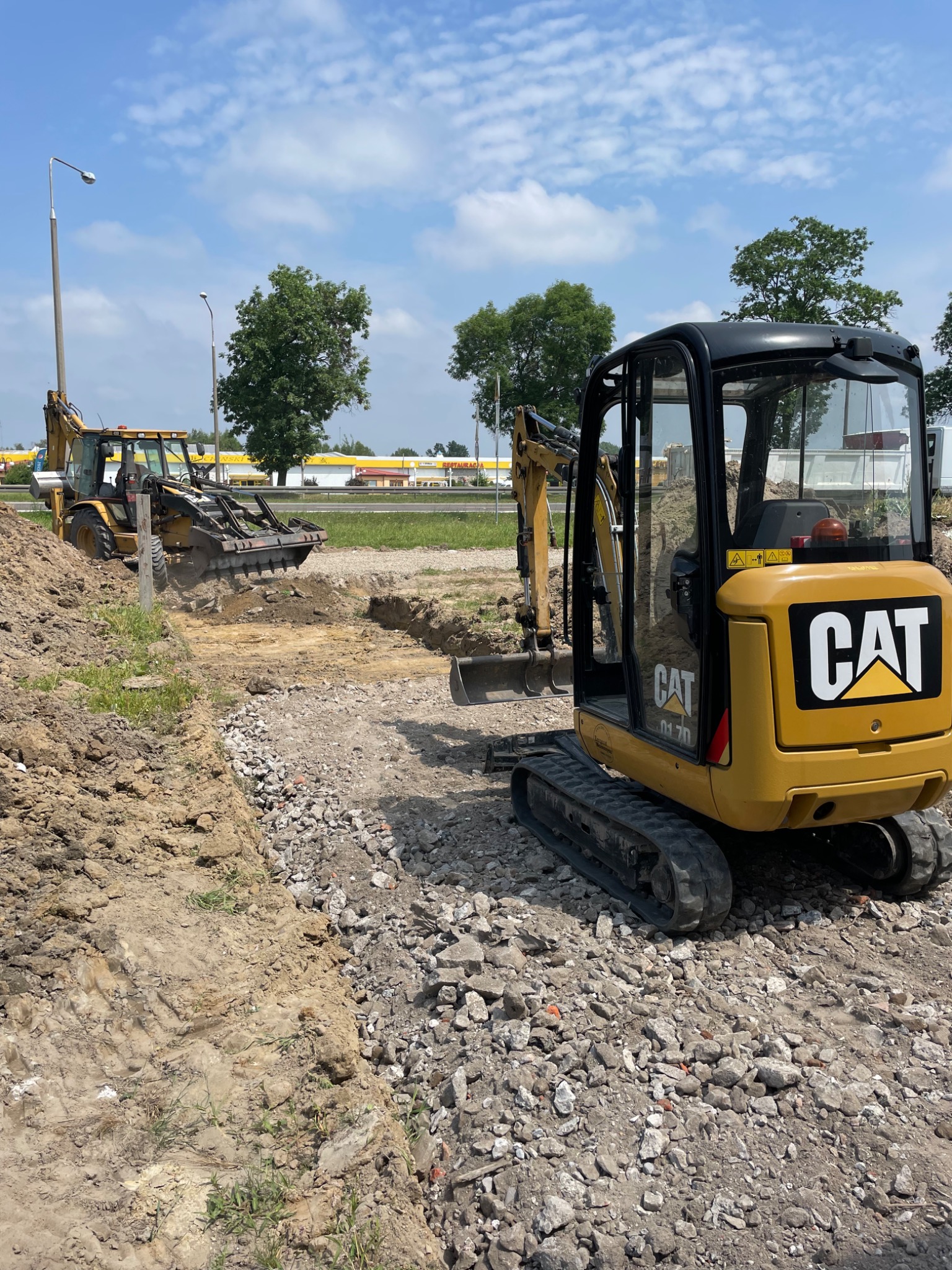 Żółta minikoparka CAT na terenie budowy z wykopanym rowem i gruzem, w tle widoczna koparko-ładowarka i budynki.