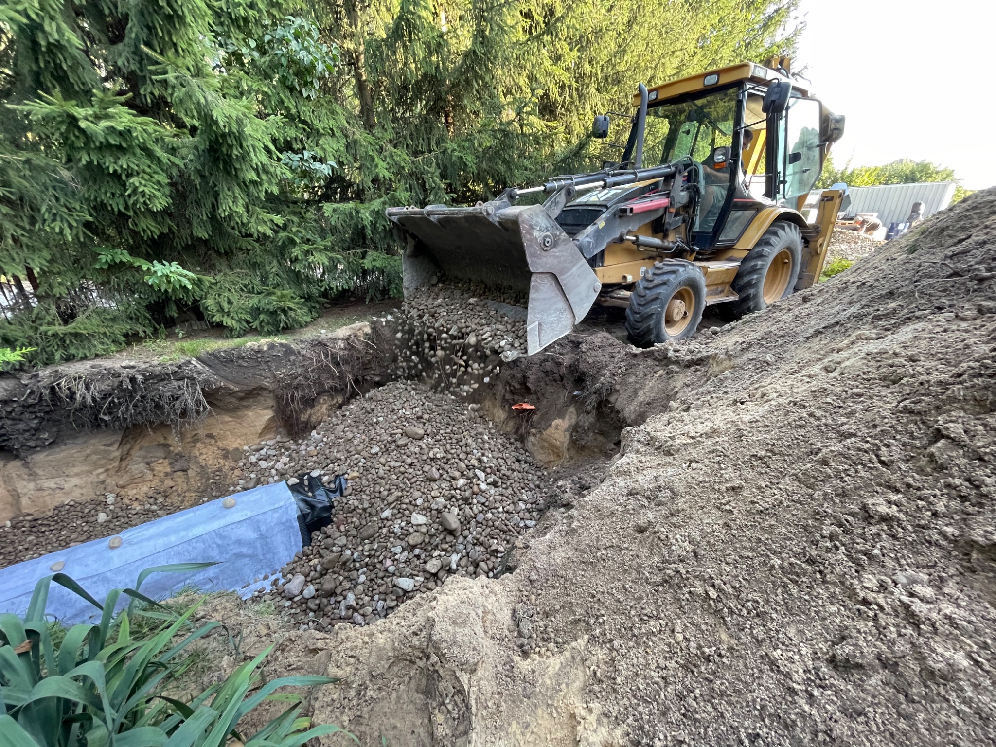Żółta koparko-ładowarka wysypuje żwir do wykopu z widocznym elementem drenażu w ziemi, na tle zielonych drzew.