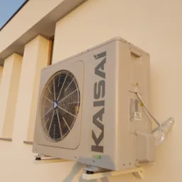 Zewnętrzna jednostka klimatyzacji Kaisai zamontowana na ścianie budynku z widocznym przewodem odprowadzającym, oświetlona popołudniowym słońcem.