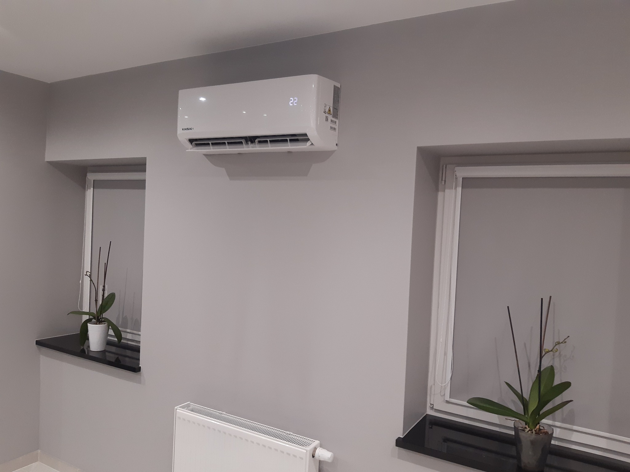 Biały klimatyzator marki Kaisai zamontowany na szarej ścianie w pomieszczeniu z dwoma oknami, na parapetach doniczki z storczykami, widoczny biały grzejnik.