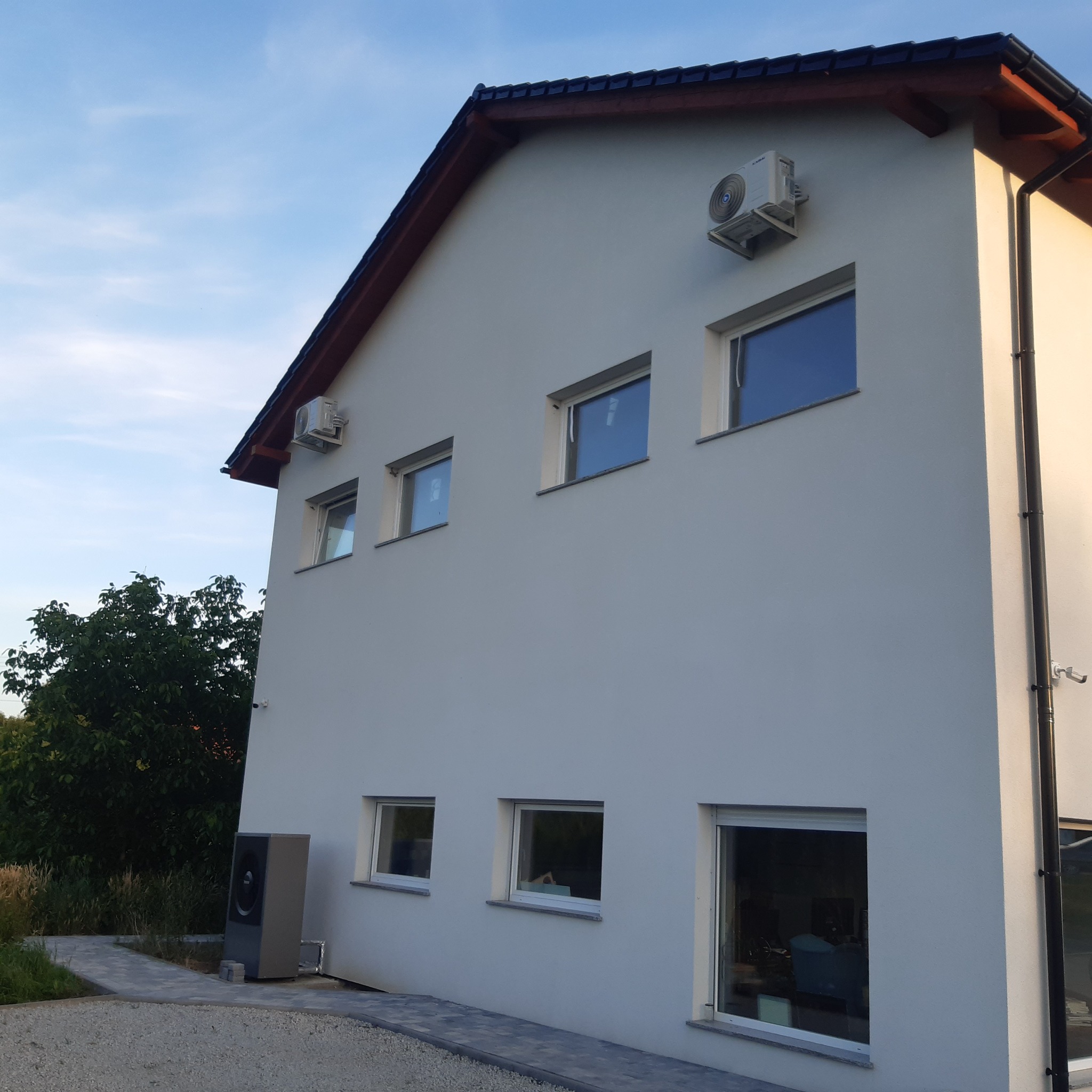 Elewacja dwupiętrowego domu z zamontowanymi klimatyzatorami na górze i pompą ciepła na dole, widok z zewnątrz w słoneczny dzień.