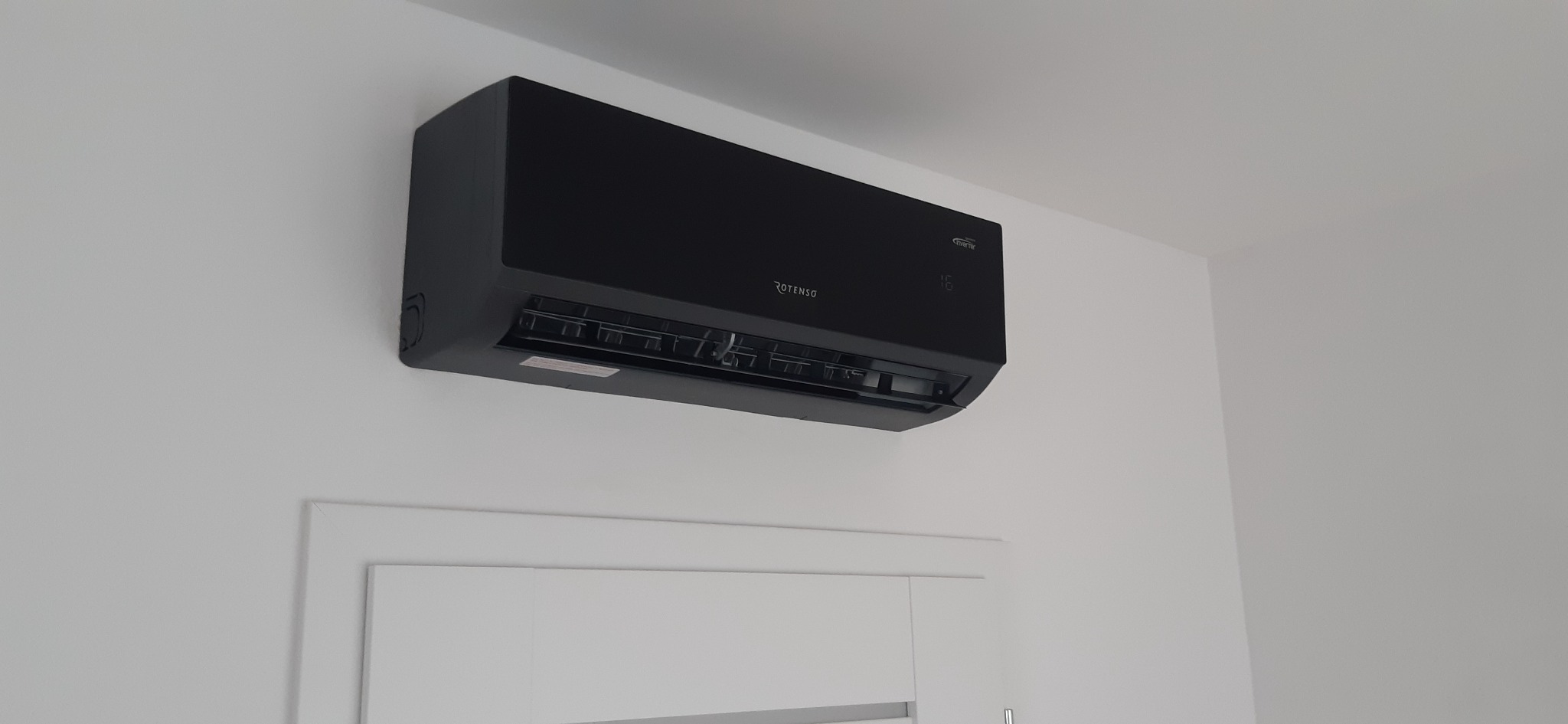 Czarna klimatyzacja Rotenso zamontowana na białej ścianie nad białą ramą drzwiową, widoczny wyświetlacz temperatury.