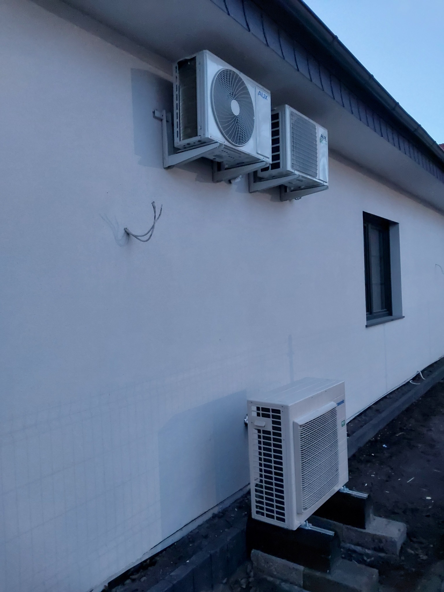 Dwa klimatyzatory marki AUX zamontowane na ścianie budynku, z wystającymi przewodami. Dodatkowo, na ziemi stoi klimatyzator marki Panasonic, umieszczony na betonowych bloczkach.