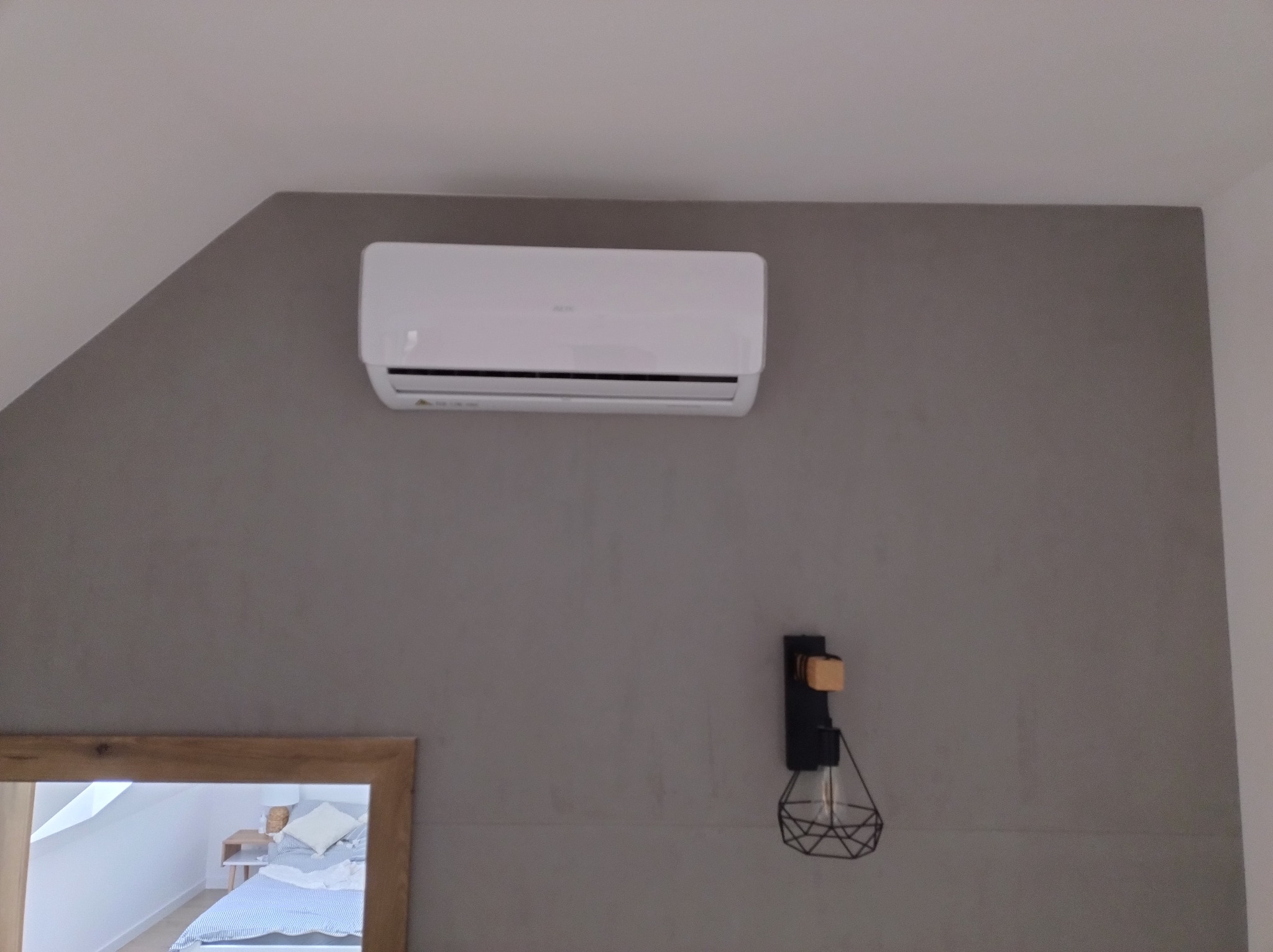 Biały klimatyzator zamontowany na szarej ścianie w sypialni, widoczny w lustrze, obok czarnej lampy ściennej z żarówką edisona.