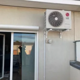 Zewnętrzna jednostka klimatyzacji LG Dual Inverter zamontowana na ścianie budynku z widocznymi wspornikami montażowymi i elementami instalacji elektrycznej; w tle przesuwne drzwi balkonowe...