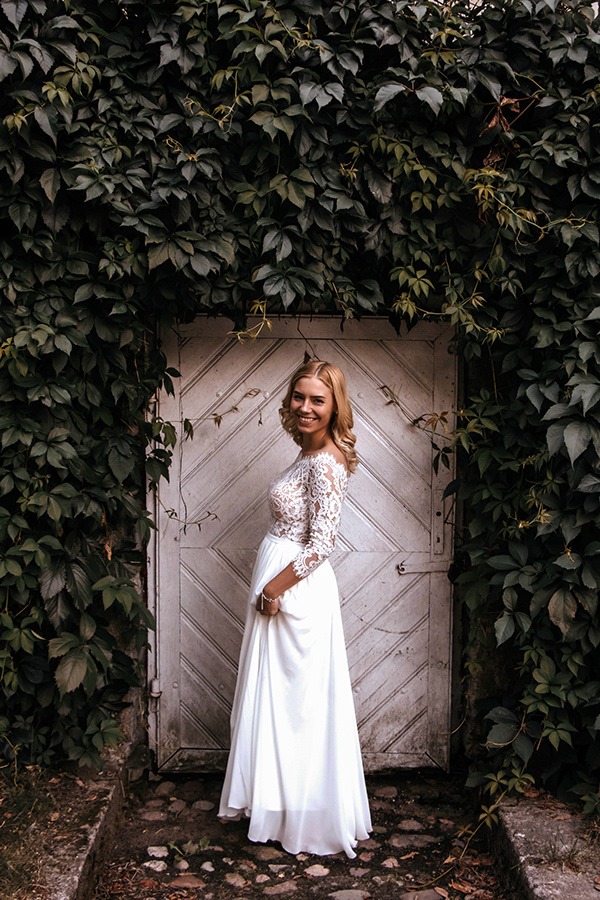 Uśmiechnięta blondynka w białej sukni z koronkową górą, pozująca na tle starej, drewnianej, pomalowanej na biało drzwi obrośniętej bluszczem.