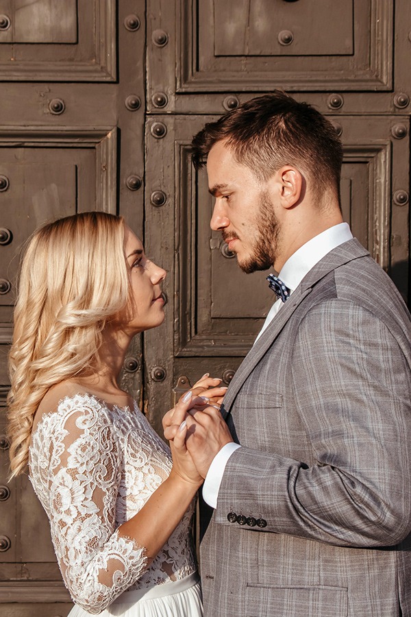 Para młoda trzymająca się za ręce na tle ciemnych, masywnych drzwi z metalowymi nitami. Panna młoda w koronkowej sukni z odkrytymi ramionami, pan młody w szarej kraciastej marynarce i muszce.