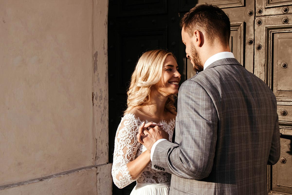 Uśmiechnięta panna młoda w koronkowej sukni i pan młody w szarej kraciastej marynarce trzymają się za ręce na tle starych, ciemnych drzwi i jasnej ściany.