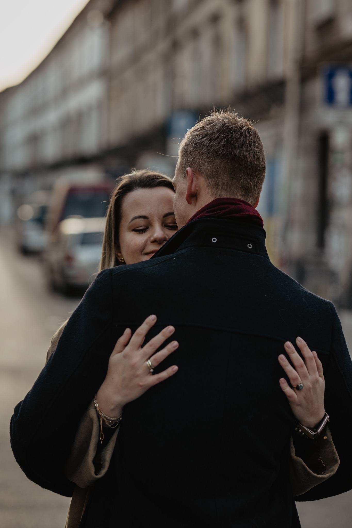 Para obejmująca się na tle miejskiej ulicy; kobieta z zamkniętymi oczami i uśmiechem, widoczne pierścionki na palcach i bransoletki na nadgarstku.