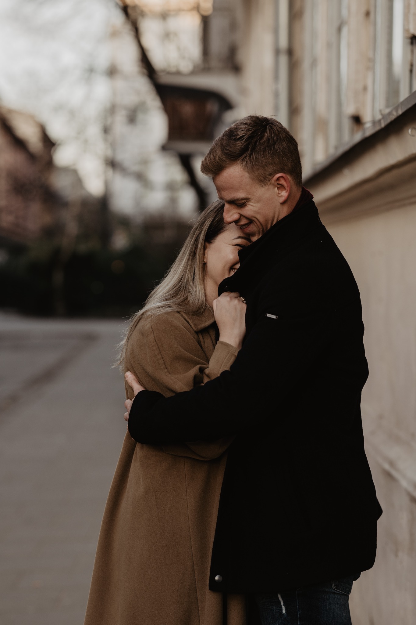 Uśmiechnięta para w objęciach na tle miejskiej zabudowy, kobieta w brązowym płaszczu, mężczyzna w czarnym, ciepłe światło.