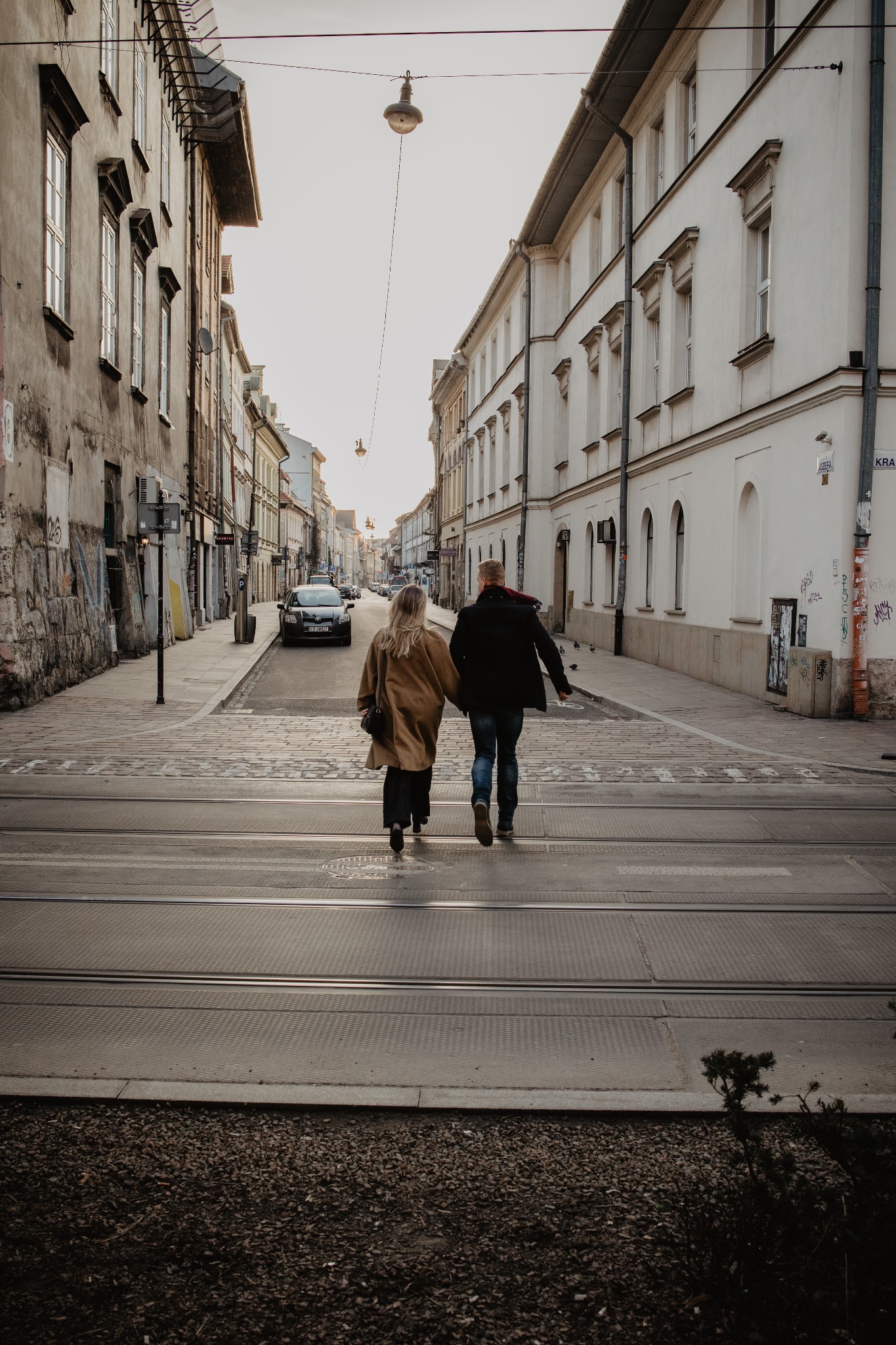 Para trzymająca się za ręce przechodzi przez tory tramwajowe na tle miejskiej ulicy z budynkami w stylu kamienic i latarniami.