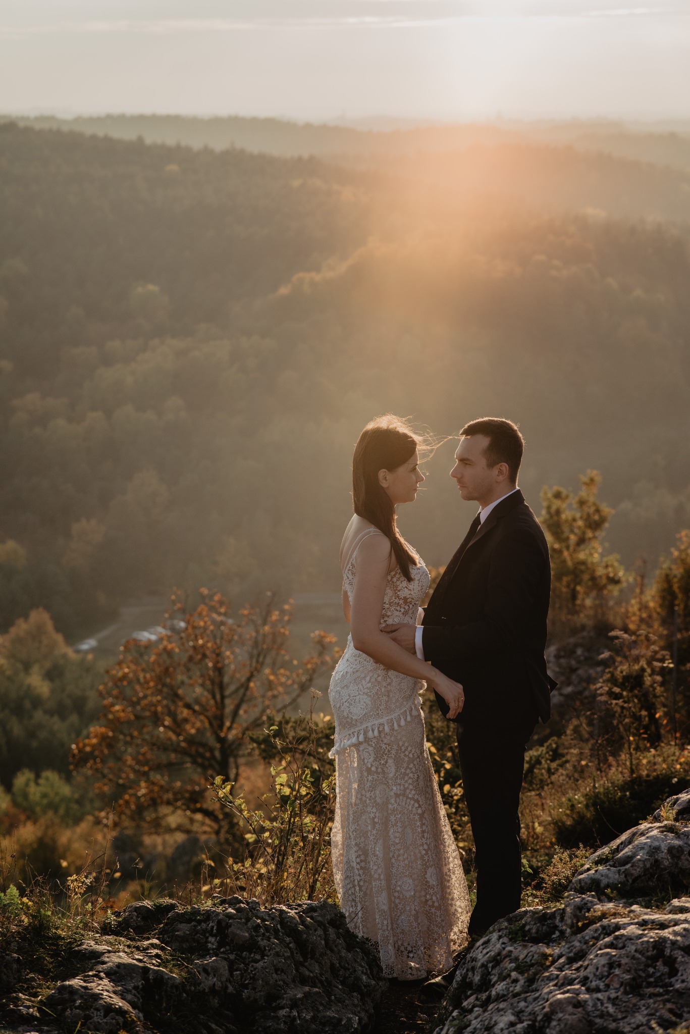 Para młoda stoi na skalistym wzgórzu, spoglądając na siebie, zalesione wzgórza i zachodzące słońce w tle.