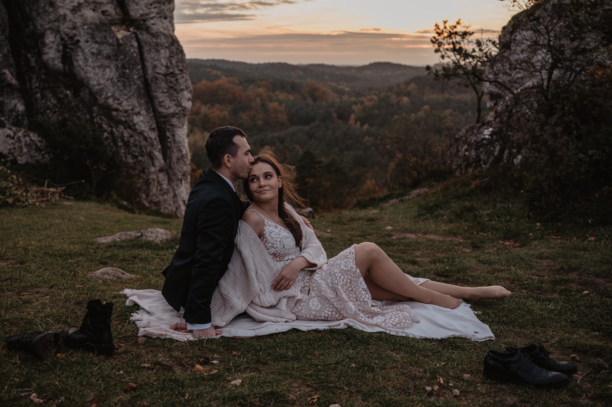 Para młoda siedzi na kocu na tle skalistego wzgórza i jesiennego lasu, mężczyzna w garniturze całuje kobietę w koronkową sukienkę w czoło, buty leżą obok.