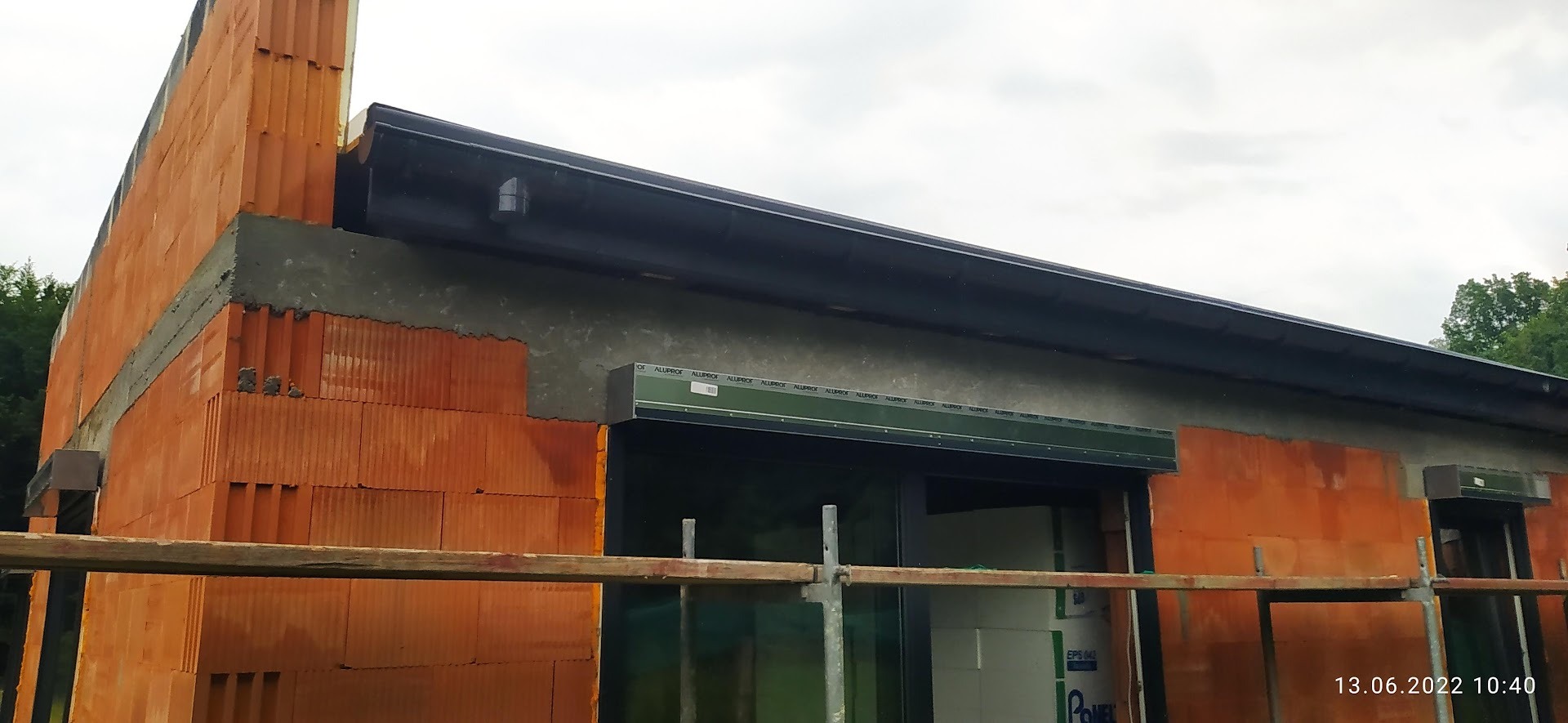 Wznoszenie ściany z pustaków ceramicznych z zamontowanymi roletami zewnętrznymi, widoczne rusztowanie i data na dole zdjęcia.