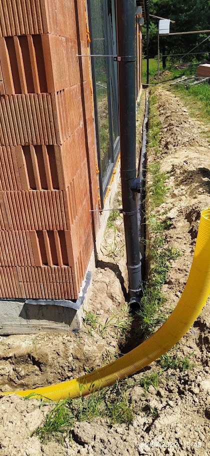 Ściana domu z pustaków ceramicznych w trakcie budowy, widoczna rynna spustowa i żółta rura drenażowa w wykopie.