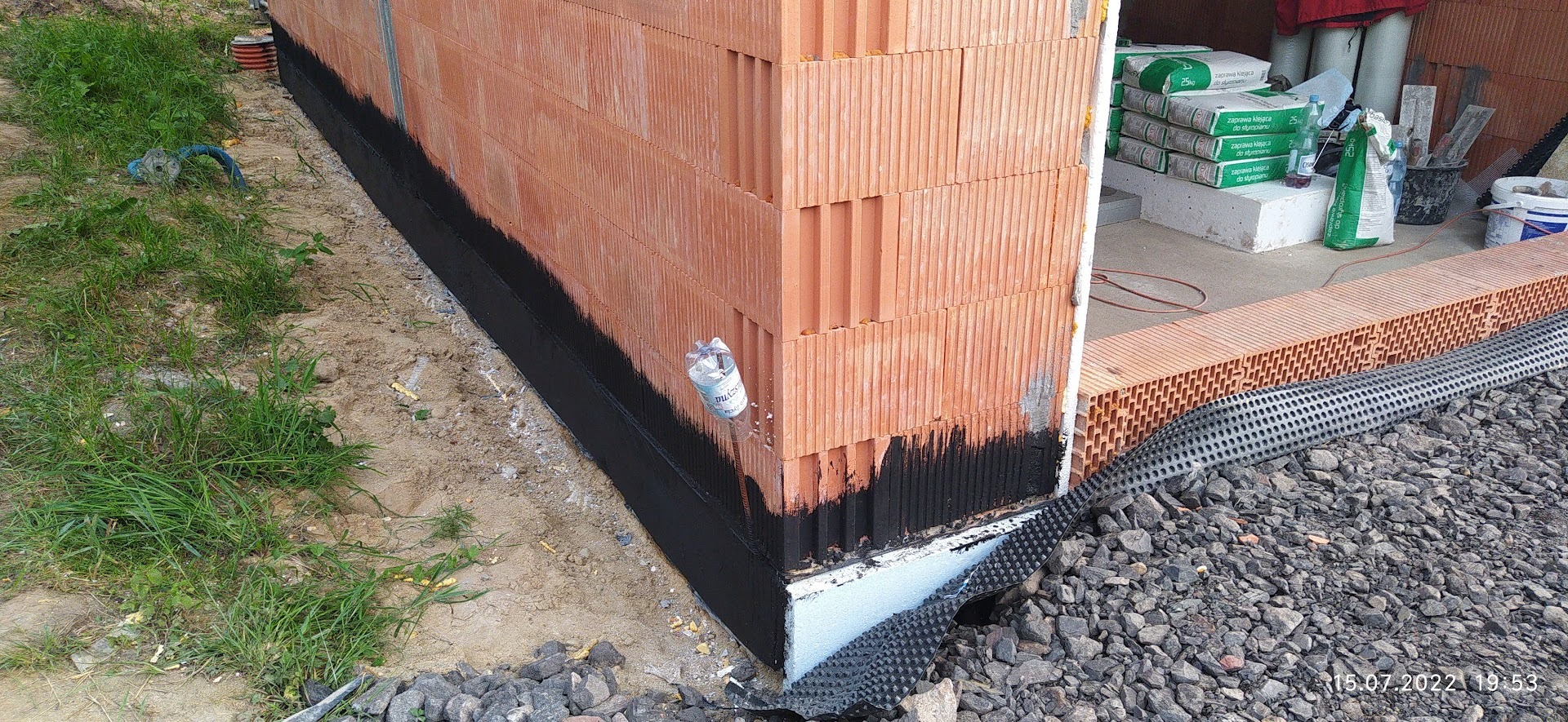 Fragment fundamentu domu z pustaków ceramicznych, zabezpieczonego izolacją pionową i folią kubełkową, z butelką wody na drucie wbitym w ziemię.