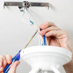 Elektryk podłącza białą oprawę oświetleniową do przewodów w suficie za pomocą śrubokręta z niebieską rączką, widoczne niebieskie i białe przewody.