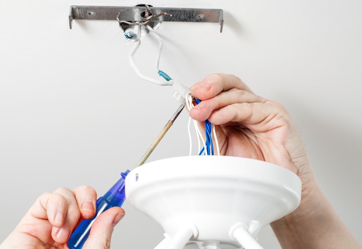 Elektryk podłącza białą oprawę oświetleniową do przewodów w suficie za pomocą śrubokręta z niebieską rączką, widoczne niebieskie i białe przewody.