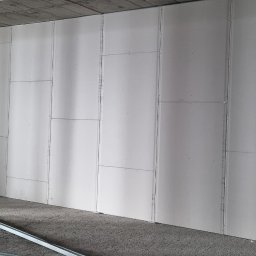 Ivan Polianskyi - Ściana z płyt gipsowo-kartonowych w surowym wnętrzu, widoczne łączenia i metalowe profile na podłodze. Szare tło sufitu i podłogi podkreśla biel ścian.