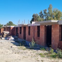 Ivan Polianskyi - Wznoszenie szeregu budynków z czerwonej cegły, widoczne otwory okienne i betonowe nadproża. Na pierwszym planie gruz i roślinność, w tle zaparkowany biały bus.