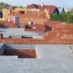 Ivan Polianskyi - Budowa domu jednorodzinnego: fundamenty z wylewką i częściowo wymurowane ściany z czerwonej cegły Porotherm. Widoczne palety z cegłami i narzędzia budowlane.
