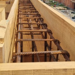 Ivan Polianskyi - Zbrojenie w drewnianym szalunku, widok z góry. Na drugim planie ceglany budynek w budowie i zielone drzewa pod błękitnym niebem z chmurami.