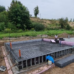 Ivan Polianskyi - Zbrojenie fundamentów w szalunku, widoczne pomarańczowe rury i różowy styropian. W tle zieleń i pochmurne niebo. Prace ziemne w toku.