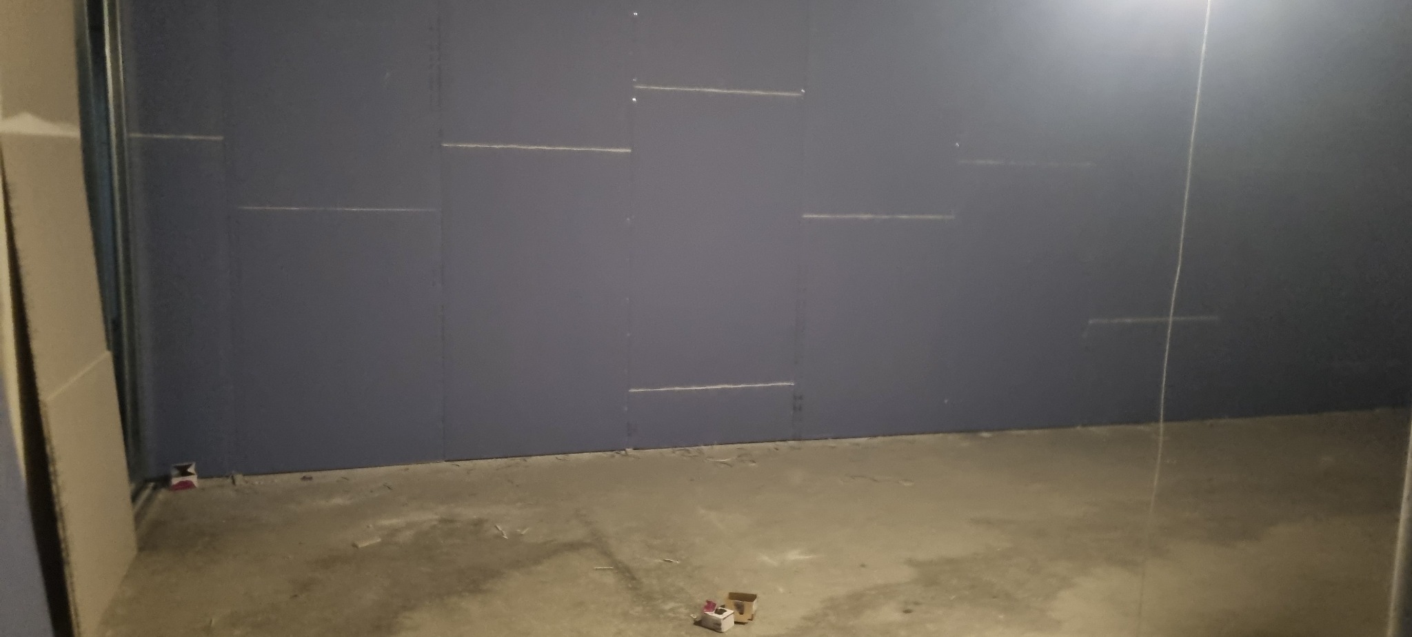 Ściana z płyt karton-gips, widoczne łączenia i szpachlowanie. Surowy beton na podłodze, fragment metalowej konstrukcji. Wnętrze w trakcie prac wykończeniowych.