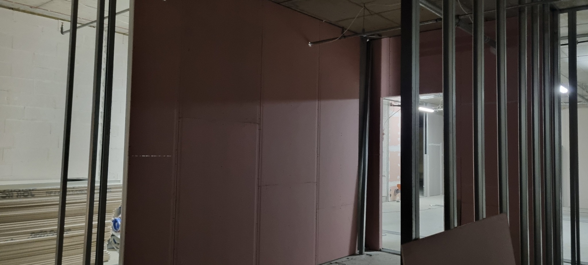 Montaż różowych płyt karton-gips na metalowym stelażu wewnątrz budynku. Widoczne kable pod sufitem i stos płyt w tle. Prace wykończeniowe w trakcie.