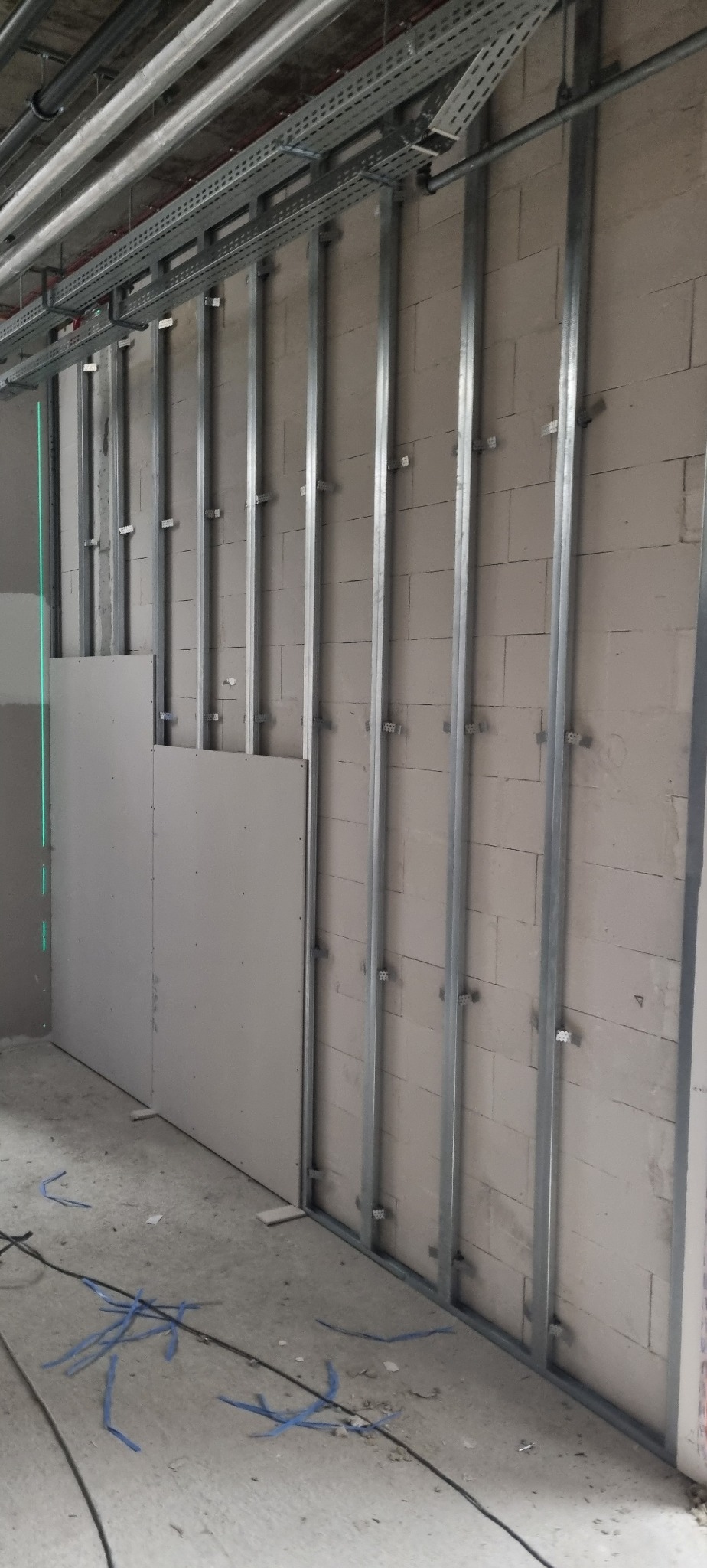 Montaż płyty gipsowo-kartonowej na metalowym stelażu przy użyciu lasera krzyżowego. Widoczne profile, fragment ściany z bloczków i przewody na podłodze.