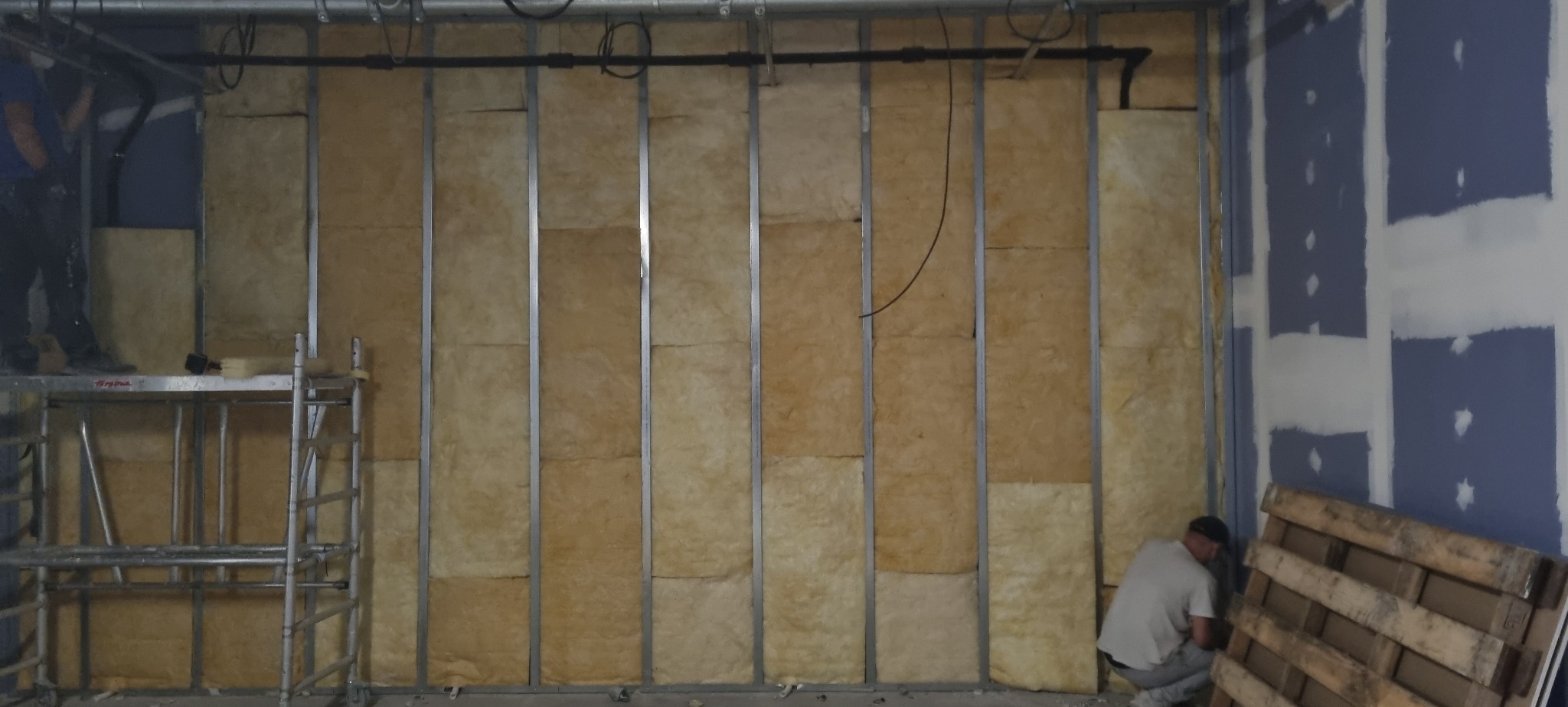 Montaż izolacji z wełny mineralnej w metalowym stelażu ściany działowej. Widoczny fragment rusztowania i pracownik w trakcie pracy.